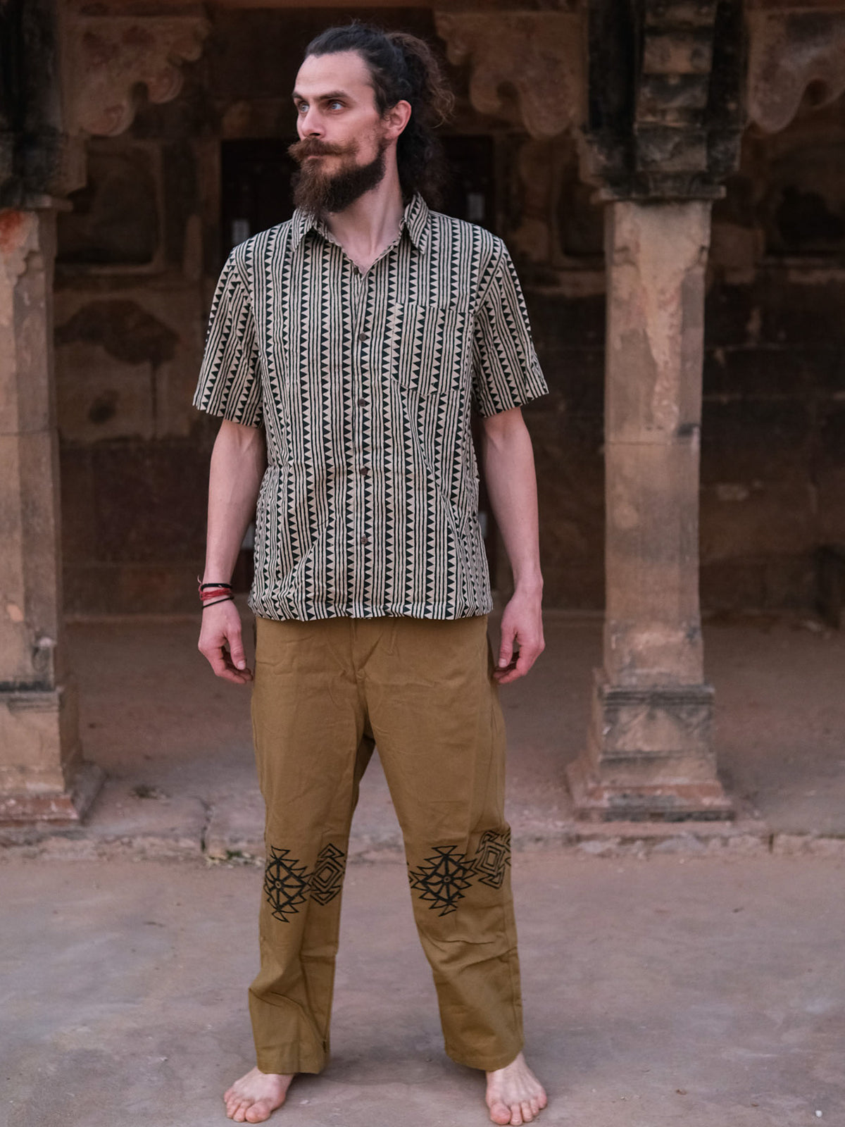 A barefoot man with a beard stands on stone ground in front of an old stone structure with arches, wearing the Tribal Triangle shirt—a unisex top featuring a vibrant Rajasthan print—paired with tan pants adorned with black designs.