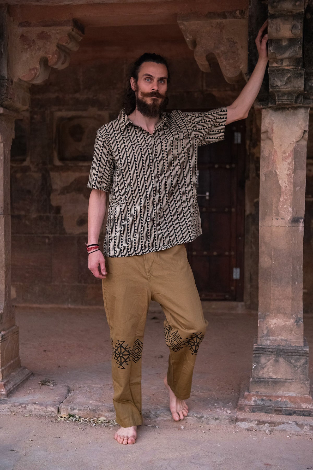 A barefoot, bearded man stands under a stone archway wearing the unisex Tribal Triangle shirt, which features a Rajasthan print and short sleeves, paired with golden-brown pants decorated with black patterns, as he rests one arm on a pillar.