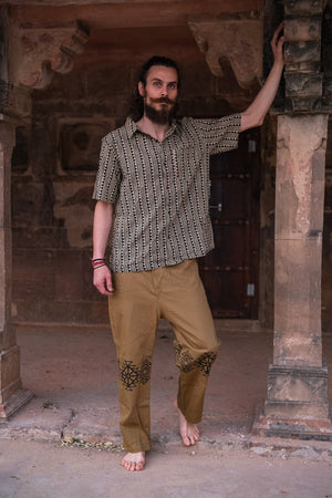 A barefoot, bearded man stands under a stone archway wearing the unisex Tribal Triangle shirt, which features a Rajasthan print and short sleeves, paired with golden-brown pants decorated with black patterns, as he rests one arm on a pillar.