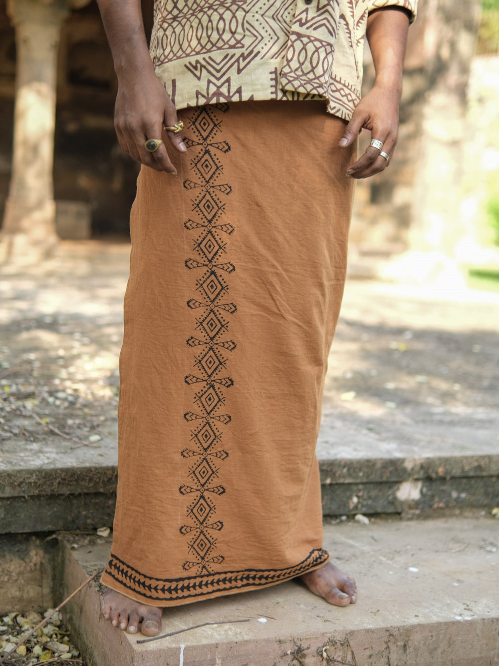 A person wearing the Dharava Tribal Cotton Dhoti in brown/black, featuring black geometric embroidery inspired by traditional block-print dhoti patterns, stands barefoot on outdoor stone steps. Only the lower half of their body is visible.