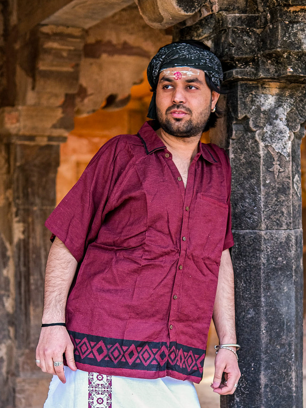 A man with a tilak on his forehead wears the maroon Atman shirt, featuring eco-friendly buttons, paired with a white dhoti as he leans against a stone pillar in a historic building showcasing classic Rajasthani architecture.