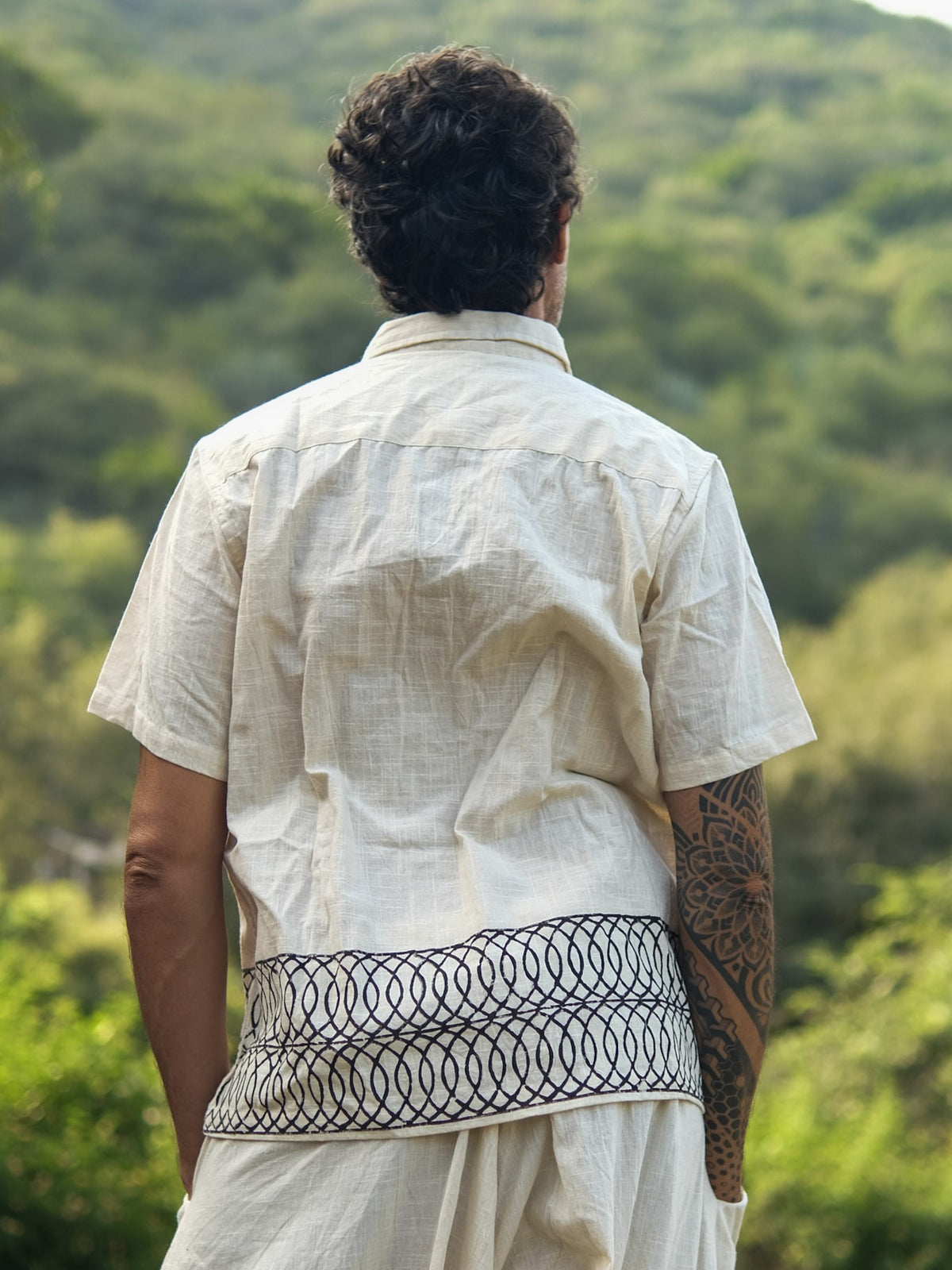 A person with dark, curly hair stands outdoors, viewed from behind, wearing the Vairagya Shirt—a light short-sleeve shirt made from organic cotton and adorned with a dark geometric pattern along the hem; lush green trees and hills fill the background, embodying a dedication to conscious fashion.