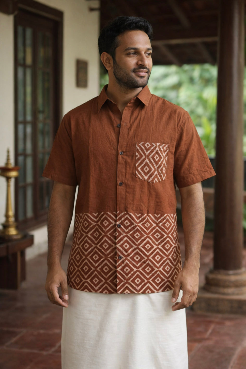 A man stands on a porch wearing the Uday Shirt in an earthy rust shade with a geometric block print, paired with a white traditional wrap. He is looking to the side and smiling softly.