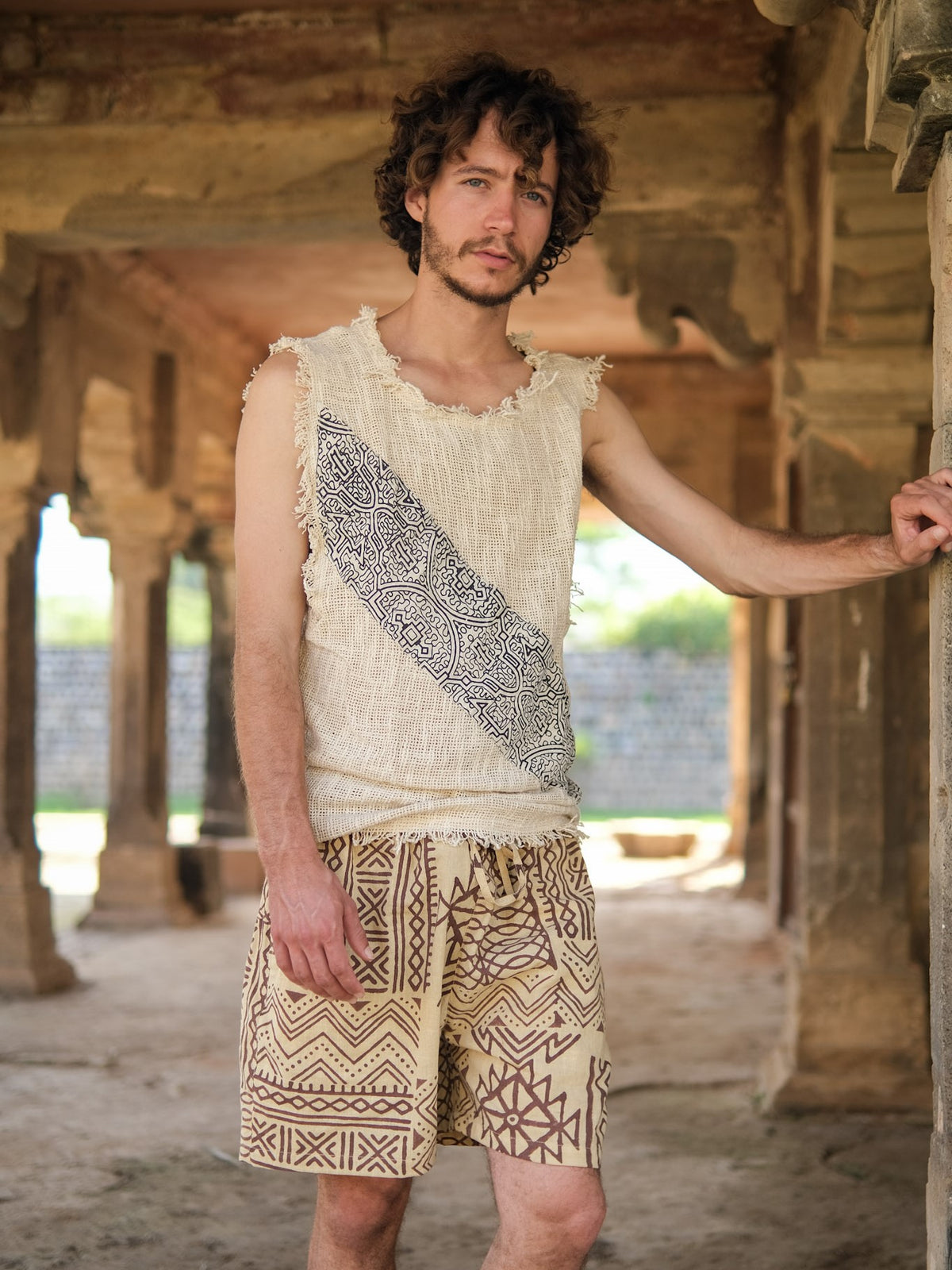 A man with curly hair, dressed in a Shipibo Tank top and patterned shorts, stands in a rustic stone structure, resting one arm on a pillar.