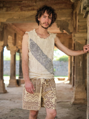 A man with curly hair, dressed in a Shipibo Tank top and patterned shorts, stands in a rustic stone structure, resting one arm on a pillar.