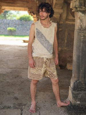 A man with curly hair stands barefoot outdoors, wearing the Shipibo Tank top—a sleeveless cream boho piece featuring a patterned stripe—paired with tan shorts adorned with tribal designs. Stone arches and lush greenery serve as the backdrop.
