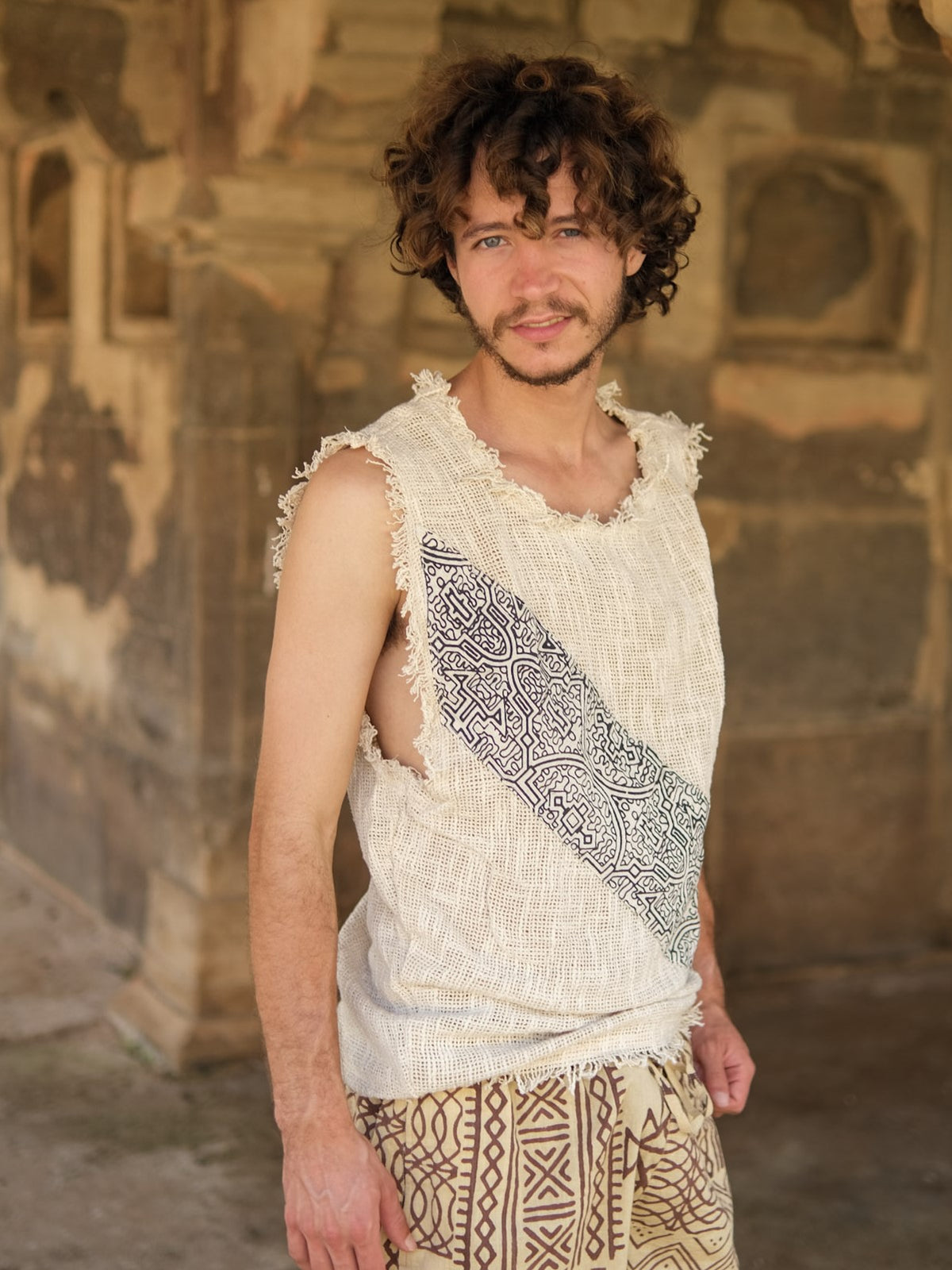 A man with curly hair and a short beard stands outdoors, wearing the Shipibo Tank top—a sleeveless, frayed beige boho piece featuring a patterned stripe—paired with matching patterned pants. Stone walls adorned with carved details form the backdrop.
