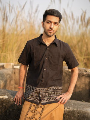 A man wearing the black Maja Shirt, a short-sleeve bohemian top with a patterned hem and subtle Rajasthani hand block print details, pairs it with a tan dhoti as he stands outdoors near a stone structure with tall grasses in the background. The artisanal accents of the shirt elevate his look.