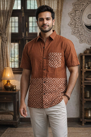 A man stands indoors wearing the Uday Shirt—an earthy rust shirt featuring white geometric block print patterns—paired with light pants. Behind him are a window, a lamp on a table, shelves, and decorative wall art.