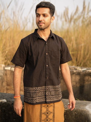 A man stands outdoors wearing the Maja Shirt in black, which features a patterned hem, and a brown wrap showcasing traditional Rajasthani hand block print. Tall grasses and a stone structure are in the background.