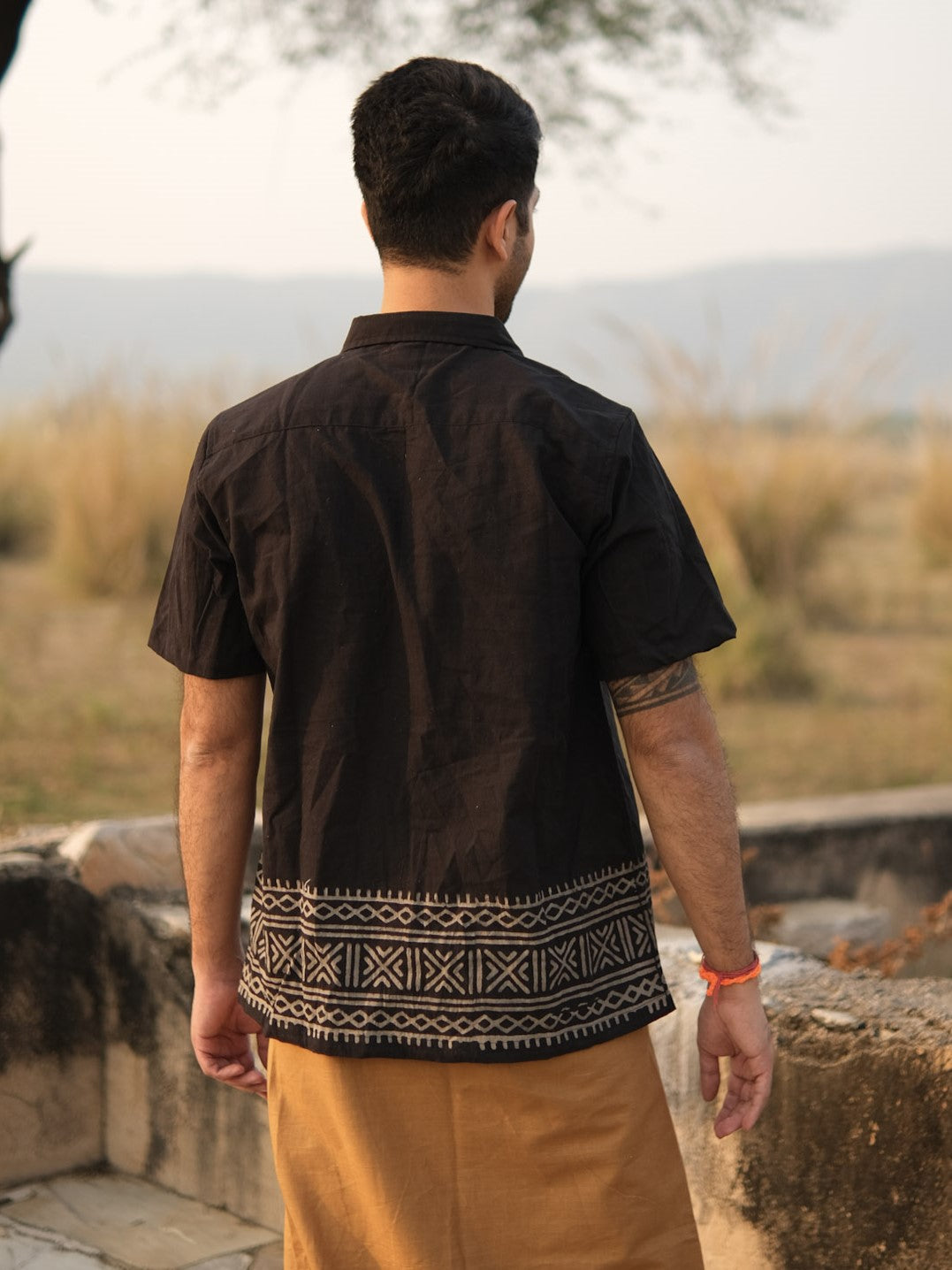 A man stands outdoors with his back to the camera, wearing the Maja Shirt—a black short-sleeve bohemian shirt featuring white geometric patterns at the bottom—paired with light brown pants. Grassy fields and hills are visible in the background.