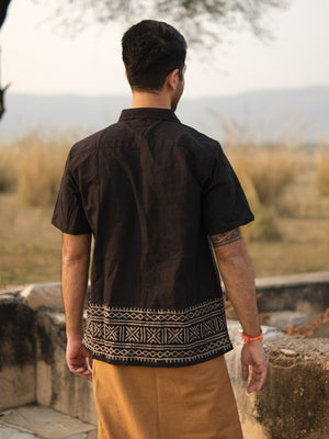 A man stands outdoors with his back to the camera, wearing the Maja Shirt—a black short-sleeve bohemian shirt featuring white geometric patterns at the bottom—paired with light brown pants. Grassy fields and hills are visible in the background.