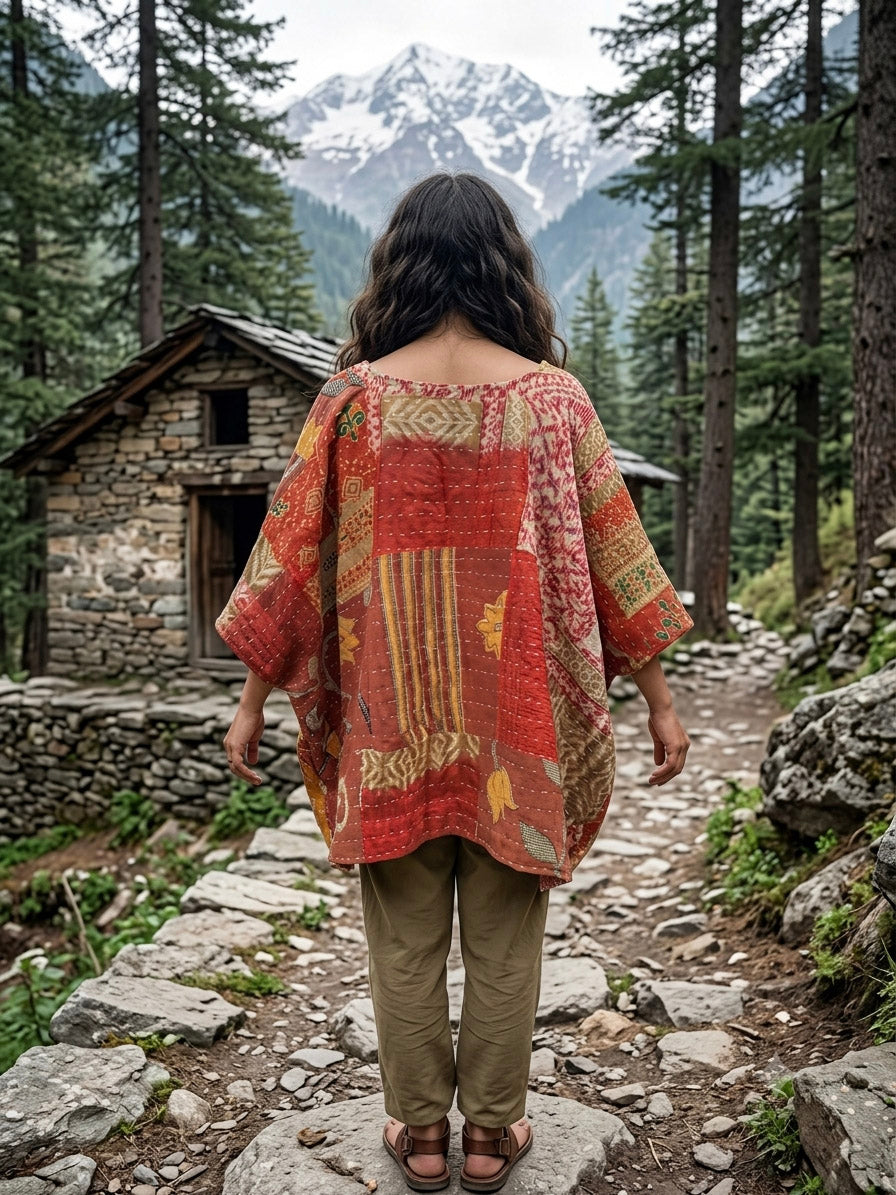 Wearing the Arohi Kantha Kaftan, a person with long hair stands on a rocky path in a forest, facing a small stone cabin with snow-capped mountains rising in the background.