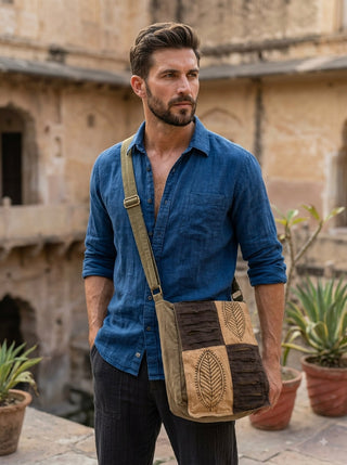 A man with short brown hair and a beard, dressed in a blue shirt and dark pants, stands outdoors near some plants. He carries the Vanika Sling Bag in dung green—a bohemian crossbody bag showcasing artisanal craftsmanship and leaf designs. An old building is visible in the background.