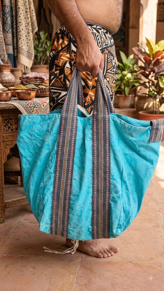 A person holds the Anoop Tote Bag—an oversized, bohemian accessory crafted from large turquoise cotton canvas with striped handles—standing barefoot on a tiled floor amidst plants and patterned textiles in the background, making it an ideal everyday shoulder bag for stylish versatility.