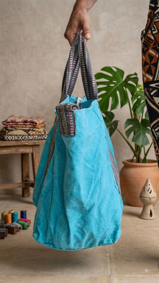 A person holds the Anoop Tote Bag, an oversized bohemian accessory in striking blue fabric with patterned handles. In the background, a plant in a clay pot, a stool with folded cloth, and colorful toy blocks rest on the floor.
