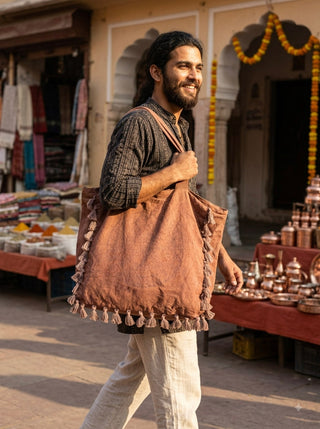 A man with long hair and a beard smiles while walking outdoors, carrying the Aatman Tassel Tote Bag adorned with hand block printed motifs. Market stalls with textiles, spices, and copperware are visible in the background.