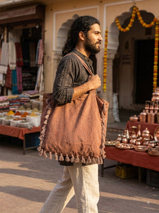 A man with long hair and a beard walks through a market carrying the Aatman Tassel Tote Bag, adorned with hand block printed motifs. Stalls display textiles, metal pots, and marigold decorations in the background.