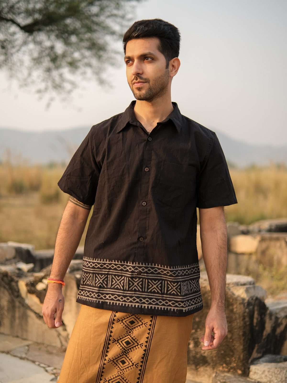 A man stands outdoors amidst a blurred natural background of grass, trees, and distant hills, wearing the Maja Shirt in black—a short-sleeve khadi cotton shirt featuring geometric Rajasthani hand block print patterns—paired with a brown garment adorned with similar designs.