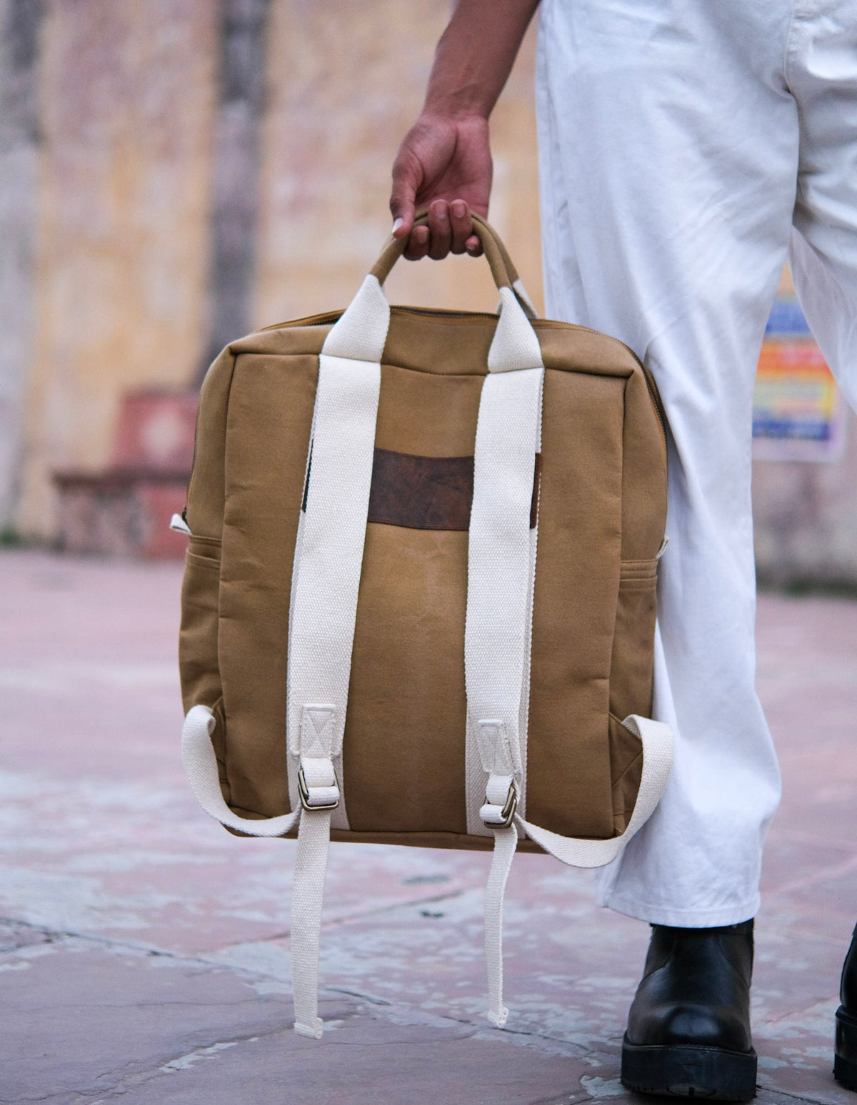 Clad in white pants and black boots, this individual readies for adventure by grasping the top handle of the Kelly Back Pack—distinguished by its brown body and white straps—while standing on a tiled outdoor surface.