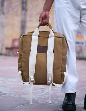 Clad in white pants and black boots, this individual readies for adventure by grasping the top handle of the Kelly Back Pack—distinguished by its brown body and white straps—while standing on a tiled outdoor surface.