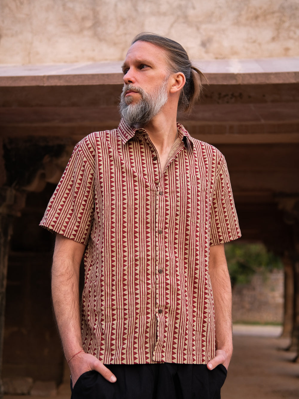 A man with a gray beard and tied-back hair stands outdoors with his hands in his pockets, looking to the side while wearing a short-sleeved, unisex Tribal Triangle shirt in red with a vibrant Rajasthan print.