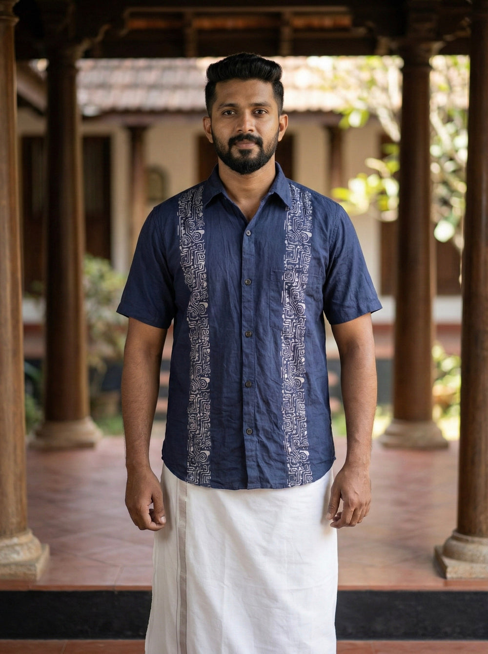 A man with short hair and a beard stands facing forward, wearing the blue Utopian Shirt and a white dhoti, on a porch with wooden pillars and a traditional house in the background.
