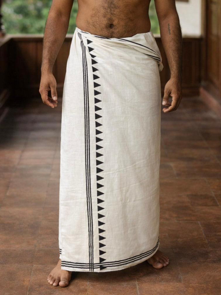 A man stands barefoot indoors on a tiled floor, wearing the Ivaan Dhoti—a handcrafted white garment featuring a black geometric border—embodying traditional style, with his upper body bare and hands resting by his sides.