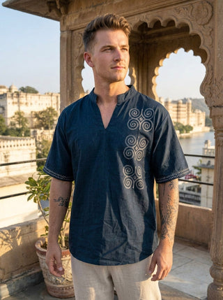 A man wearing the Midnight Nomad kurta, a black shirt adorned with white embroidered patterns inspired by contemporary nomadic fashion, stands on a terrace with historic buildings and water in the background, gazing off to the side.