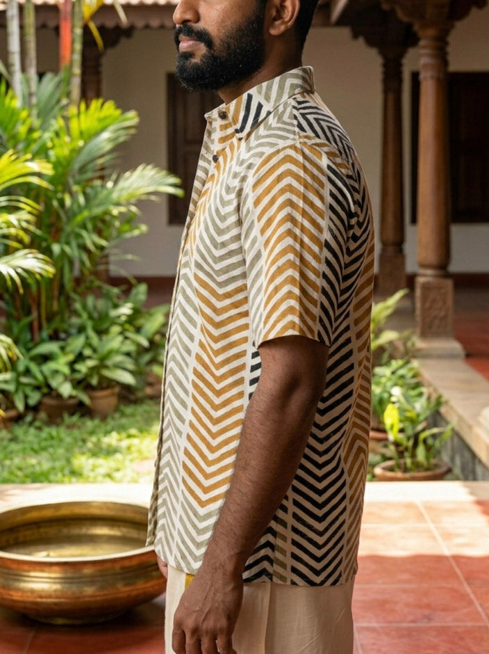 A man stands sideways outdoors near green plants and a brass bowl on a tiled patio, wearing the Taar Chevron Shirt—a relaxed fit shirt featuring a brown, yellow, and black chevron pattern.