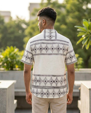 A man stands outdoors with his back to the camera, wearing the Adiva Shirt—crafted from white cotton slub and adorned with black tribal geometric detailing—paired with light-colored pants. Green trees and blurred buildings in the background create a relaxed, resort wear ambiance.