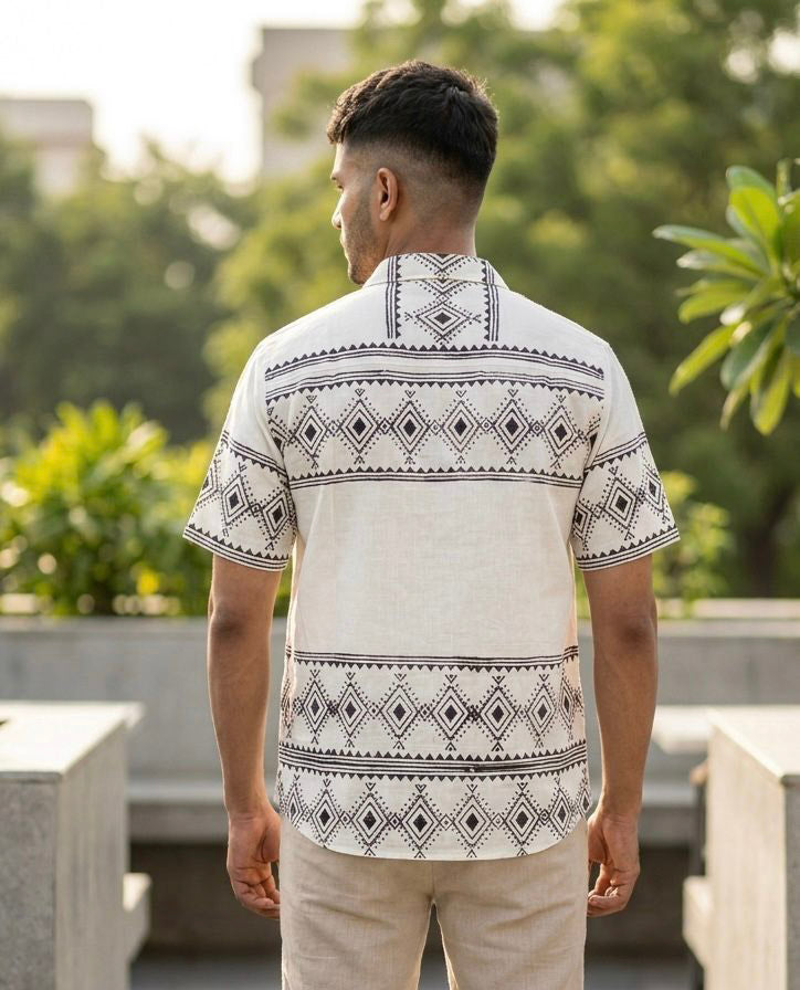 A man stands outdoors with his back to the camera, wearing the Adiva Shirt—crafted from white cotton slub and adorned with black tribal geometric detailing—paired with light-colored pants. Green trees and blurred buildings in the background create a relaxed, resort wear ambiance.