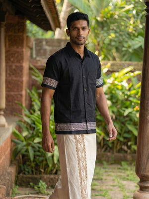 A man wearing the Adhir Shirt, an ethnic contemporary black cotton shirt with patterned borders, and a cream traditional dhoti stands outdoors near a building, surrounded by greenery.