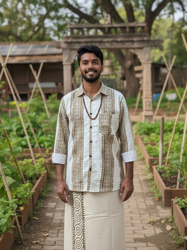 A man wearing the Dwara Shirt, an artisanal block print cotton shirt, and a traditional white dhoti stands smiling in a garden with raised plant beds, lush greenery, trees, and a stone archway in the background.