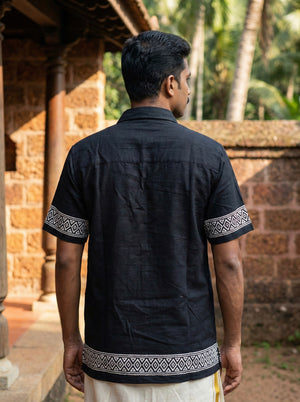 A man stands outdoors with his back to the camera, wearing the Adhir Shirt—a black cotton shirt featuring patterned trim on the sleeves and hem for an ethnic contemporary look. The background includes stone walls, pillars, and green trees.