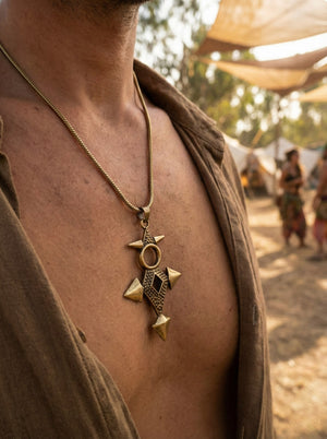 A person wearing a brown shirt open at the chest displays a large, ornate Scroll Pendant necklace. In the blurred background, people and tents are visible outdoors.