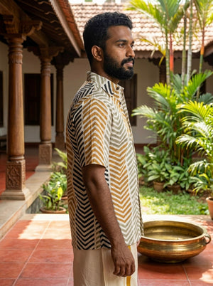 A man with a beard stands sideways on a tiled veranda, wearing the Taar Chevron Shirt—a relaxed-fit, patterned short-sleeve shirt—paired with cream pants. Lush green plants and traditional wooden pillars are in the background.