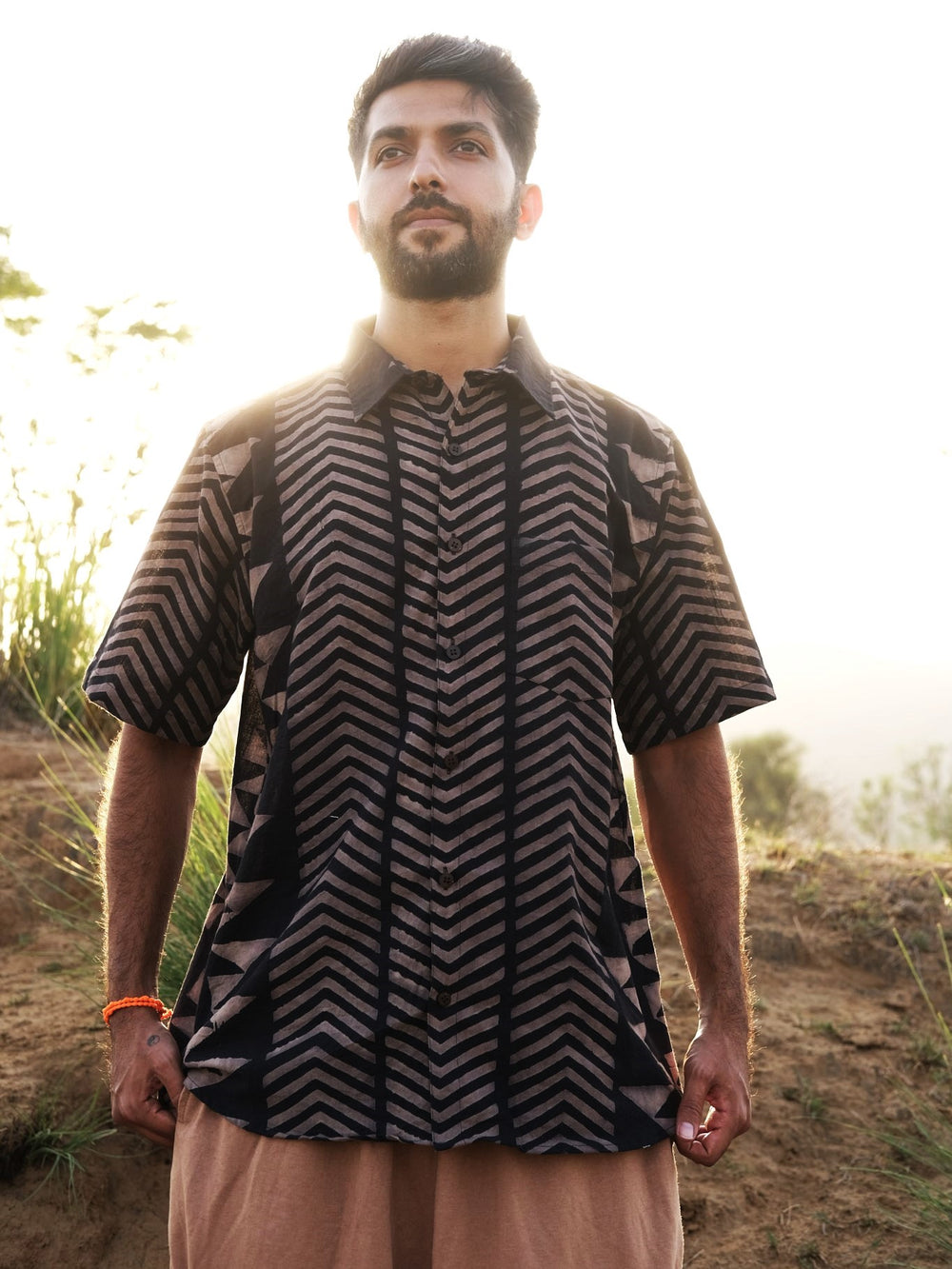 A man with a beard stands outdoors in sunlight, wearing the black Zorik Shirt and tan pants, with greenery and hills in the background.