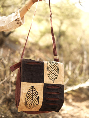 A hand holds the Vanika Sling Bag—a brown and beige patchwork bohemian crossbody with leaf patterns on the flap—outdoors in a sunlit, blurred natural setting.