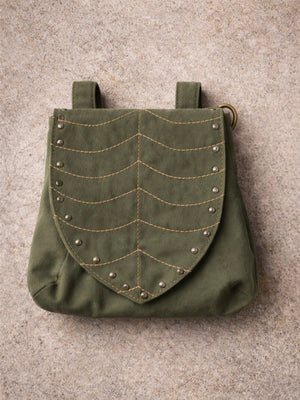 The Luna Festival Bag is an olive green fabric pouch with a leaf-flap design, detailed chevron stitching, and metal rivets, shown lying flat on a textured, light-colored surface.