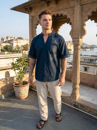 A man stands on a balcony in sunlight, dressed in contemporary nomadic fashion—a navy blue Midnight Nomad kurta with white handblock prints, paired with beige pants and sandals. Ornate arches, a potted plant, and a cityscape with buildings and water are visible in the background.