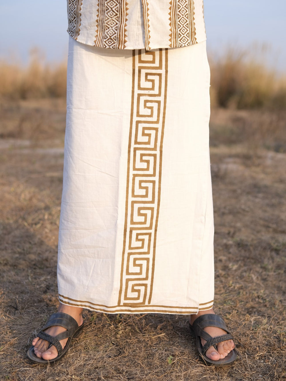 A person stands outdoors on dry grass, wearing the Rivan Dhoti in white and brown, featuring a handcrafted ethnic design with a geometric tribal border print, paired with black sandals. Only the lower half of their body is visible.