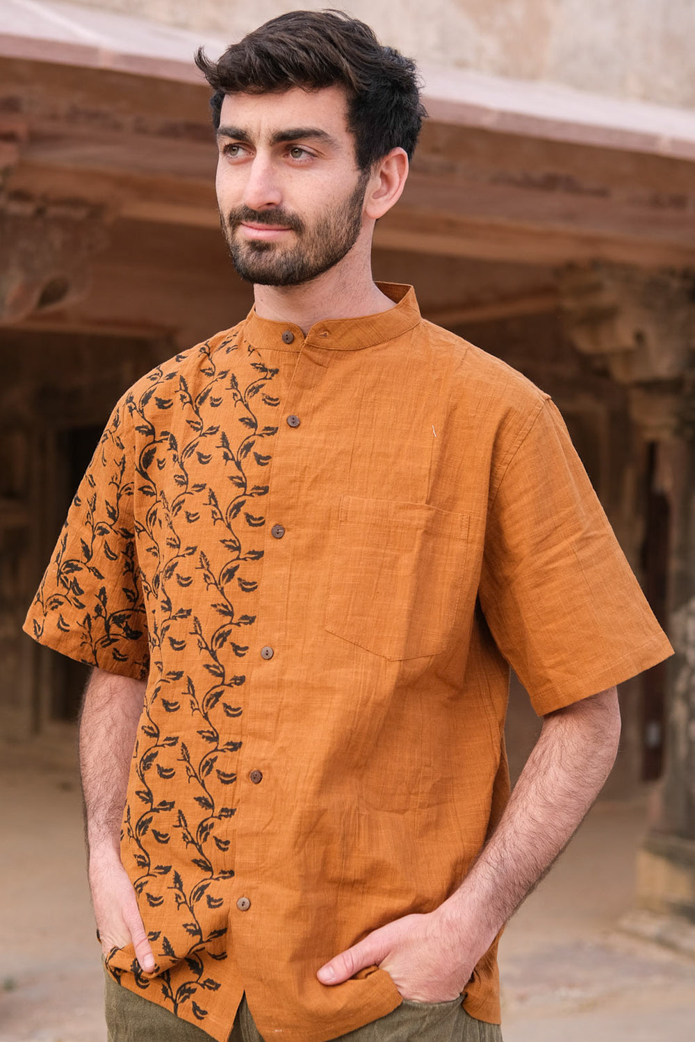 A man with short dark hair and a beard wears a brown Kunal Shirt with a leafy pattern on one side, standing outside an old rustic building. With hands in his pockets, he makes the Kunal Shirt a true statement piece.