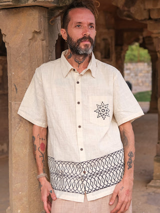 A tattooed man stands in a historic stone structure wearing a beige Vairagya Shirt made from organic cotton, featuring geometric designs on the pocket and hem. Bearded, he looks aside, with blurred stone pillars and walls framing the background.