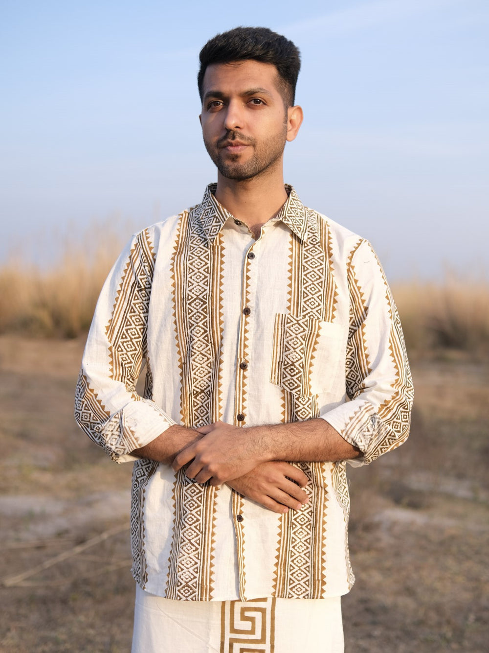 A man stands outdoors against a backdrop of dry grass and a clear sky, wearing the Dwara Shirt—a block print cotton shirt in beige and brown patterns—paired with white pants. He has short hair, a trimmed beard, and his hands are folded in front of him.
