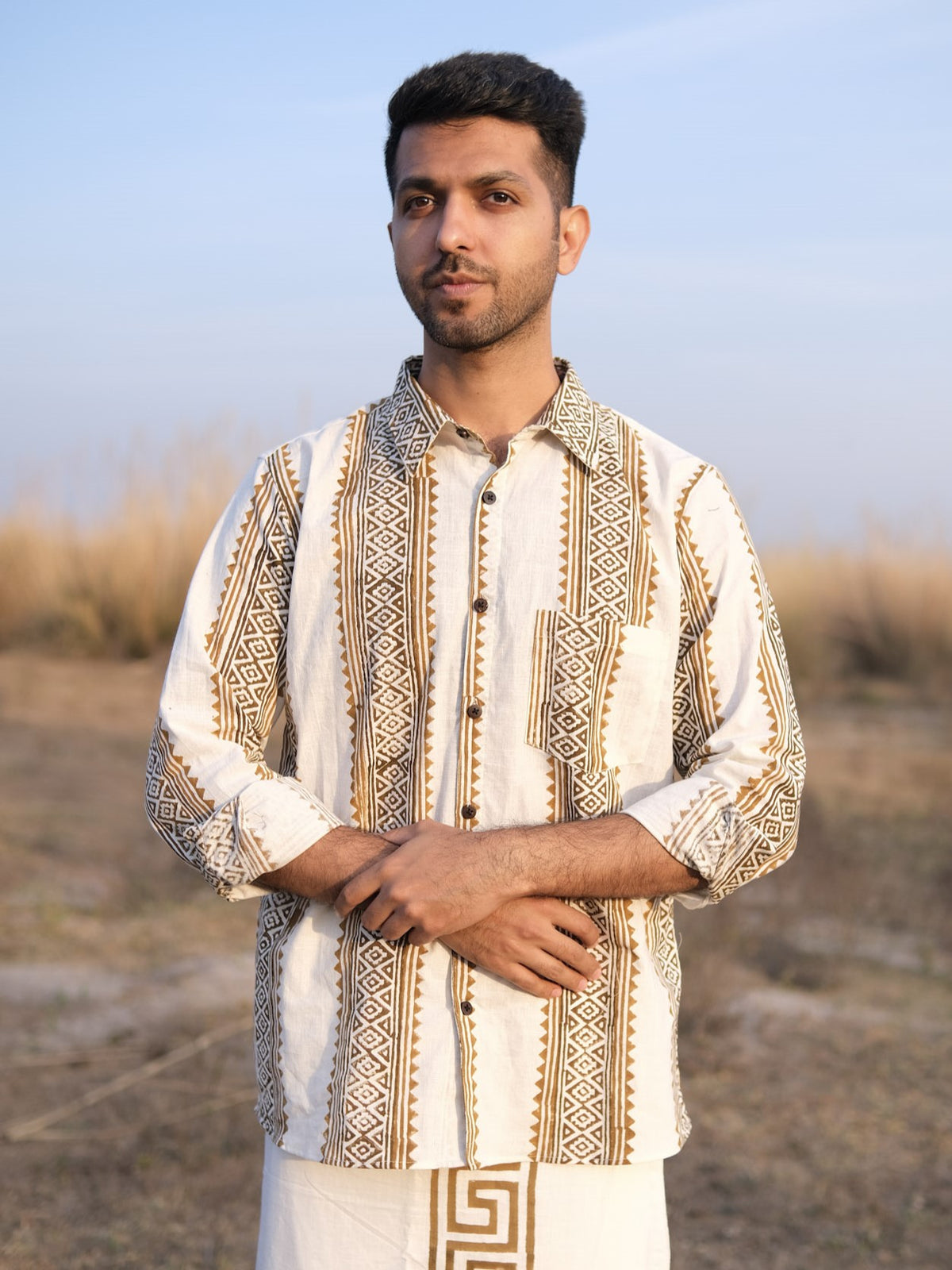 A man stands outdoors against a backdrop of dry grass and a clear sky, wearing the Dwara Shirt—a block print cotton shirt in beige and brown patterns—paired with white pants. He has short hair, a trimmed beard, and his hands are folded in front of him.