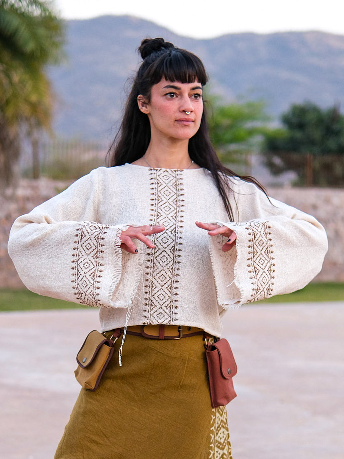 A woman stands outdoors with her hands raised, wearing the Kapisha Top—a loose, patterned white boho blouse crafted from sustainable fabric—paired with a brown skirt featuring pouches. Mountains and greenery fill the background.