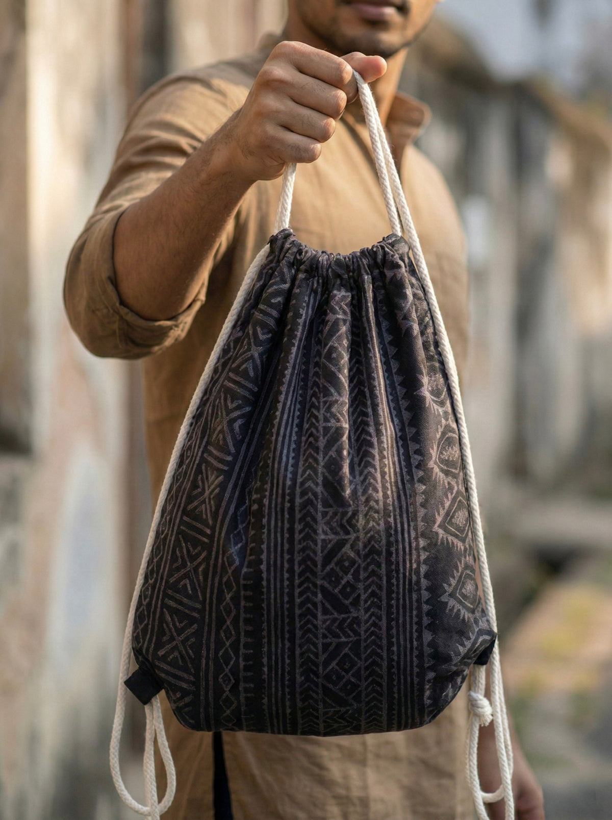 A person holds up a dark Sahaz Drawstring Bag featuring a repeating tribal pattern and white rope straps.