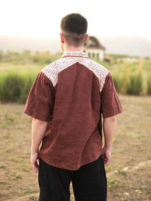 A man stands outdoors with his back turned, wearing the Rustwood Shirt—a short-sleeve brown tribal print shirt featuring a white patterned yoke. In the blurred background, grass, an old building, and distant mountains are visible.