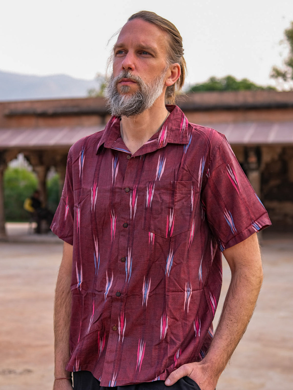A man with a beard and long hair is seen outdoors, with a building and trees in the background, wearing the maroon short-sleeved handcrafted Ikat Shirt.